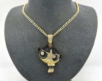May include: Gold chain necklace with a pendant featuring a cartoon character with black hair, green eyes, and a white and black outfit. The pendant is covered in clear and black cubic zirconia stones.
