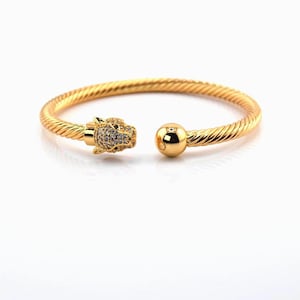 May include: A gold-colored cuff bracelet with a twisted rope design. One end features a leopard head with small, sparkling stones, while the other end has a smooth, round ball. The bracelet is displayed on a white background.