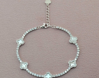 May include: A silver bracelet with clear, round gemstones and four white clover-shaped accents. The bracelet has an adjustable chain and a heart-shaped charm. The bracelet is on a pink background.