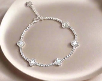 May include: A silver bracelet with a delicate design. The bracelet features a series of small, clear stones and five clover-shaped charms with a mother-of-pearl appearance. The bracelet is displayed on a light-colored, round plate.