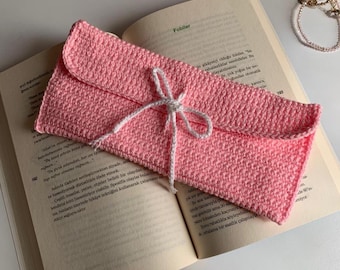 Peut inclure: Une pochette rose en crochet, de style enveloppe, attachée avec un nœud en ficelle blanche. L'étui est ouvert, révélant l'intérieur. La pochette est posée sur un livre ouvert avec du texte visible.