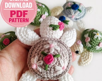 May include: A selection of handmade crochet turtle ornaments in various colours. The turtles have white, grey, and green shells, with embroidered floral details in pink and white. The image includes the text "DIGITAL DOWNLOAD PDF PATTERN".
