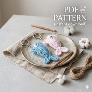 May include: Two crocheted dolphin keychains, one blue and one pink, rest on a speckled plate with a linen napkin. The keychains have black eyes and silver key rings. The image includes the text "PDF PATTERN (instant download)".