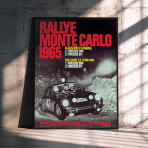 May include: A framed poster advertising the 1965 Monte Carlo Rally, featuring a Porsche race car. The poster has red text, including "RALLYE MONTE CARLO 1965" and race results. The Porsche logo is at the bottom, set against a dark background.