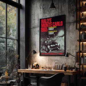 May include: A framed poster with the text "Rallye Monte Carlo 1965" in red and white, featuring a black Porsche race car. The poster is displayed on a concrete wall above a wooden desk with a black chair.