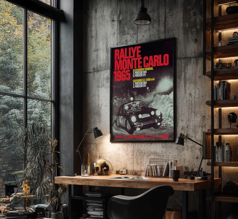 May include: A framed poster with the text "Rallye Monte Carlo 1965" in red and white, featuring a black Porsche race car. The poster is displayed on a concrete wall above a wooden desk with a black chair.