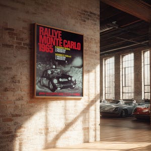 May include: A framed poster with the text "Rallye Monte Carlo 1965" in red and white. The poster features a black Porsche car and is displayed on a brick wall. The image is in a room with several classic cars.