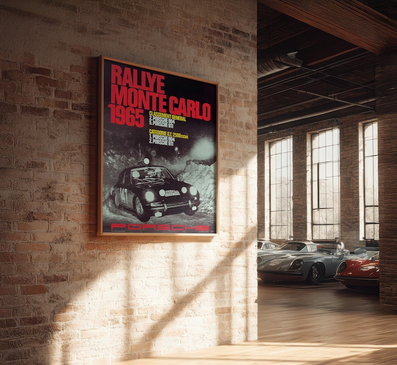 May include: A framed poster with the text "Rallye Monte Carlo 1965" in red and white. The poster features a black Porsche car and is displayed on a brick wall. The image is in a room with several classic cars.