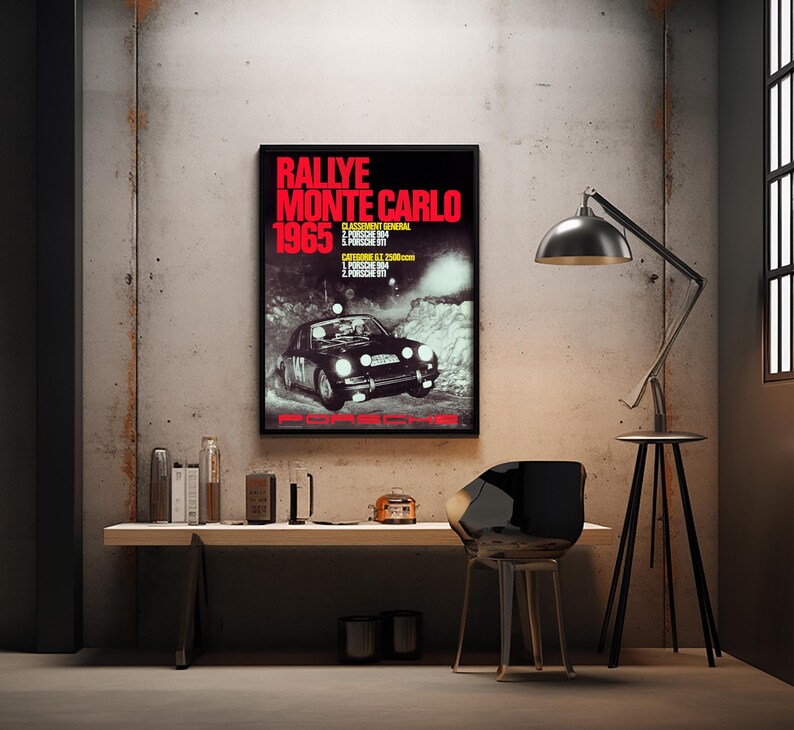 May include: A framed poster advertising the 1965 Monte Carlo Rally. The poster, with a black background and red text, shows a vintage racing car. The poster is mounted on a concrete wall, with a desk and chair in front.