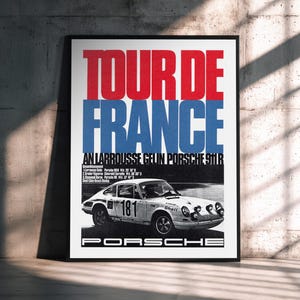 May include: A framed poster with the words "TOUR DE FRANCE" in red and blue block letters. Below the text is a black and white image of a Porsche 911R race car with the number 181. The poster is leaning against a concrete wall.