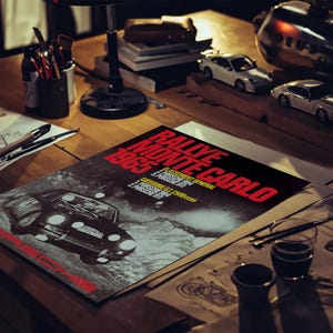 May include: A poster for the 1965 Rallye Monte Carlo with a black Porsche car illustration. The poster is on a wooden desk with model cars, art supplies, and a lamp. The poster's text is in red and white.