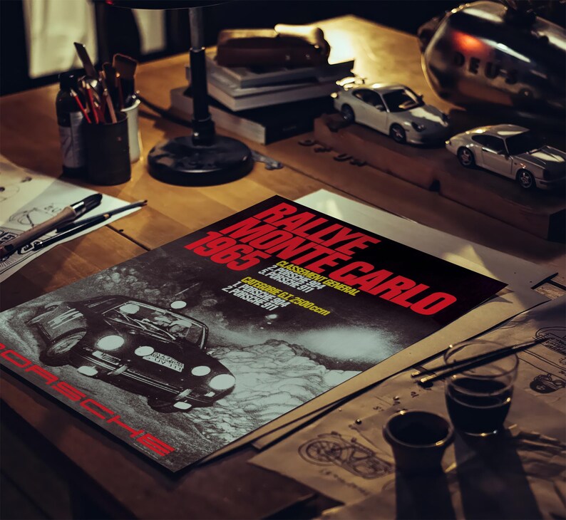 May include: A poster for the 1965 Rallye Monte Carlo with a black Porsche car illustration. The poster is on a wooden desk with model cars, art supplies, and a lamp. The poster's text is in red and white.