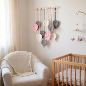 Peut inclure: Décoration murale avec des feuilles décoratives roses, grises et crème. Les feuilles sont suspendues à une tringle en bois avec des cordes. L'ensemble est dans une chambre d'enfant, avec un lit bébé et un fauteuil blanc.