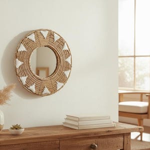 Puede incluir: Un espejo redondo con un marco de fibra natural tejida y detalles triangulares blancos cuelga de una pared blanca. Una consola de madera exhibe un jarrón con flores secas, una pequeña suculenta y una pila de libros. La habitación tiene una estética neutra y bohemia.