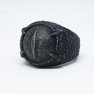 May include: A black metal ring with a textured design. The ring features a large, round, raised center piece with a detailed design.