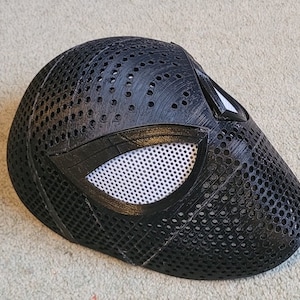 May include: A black plastic mask with a white mesh eye opening and a black spider web design. The mask is made of a 3D printed material and has a textured surface.
