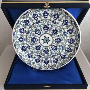 May include: A white ceramic plate with a blue and green floral design. The plate is in a blue velvet box.