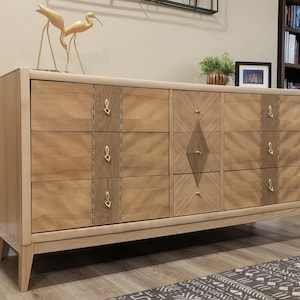 May include: A light wood chest of drawers featuring nine drawers with gold-toned hardware. The central section displays a diamond-shaped inlay pattern, while the side sections feature vertical textured wood panels. The piece stands on four tapered legs.