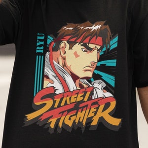 Puede incluir: Camiseta negra con un gráfico de Ryu de Street Fighter. El gráfico está en un estilo retro con un esquema de color rojo, amarillo y azul. El texto "RYU" y "STREET FIGHTER" está impreso en la camiseta.