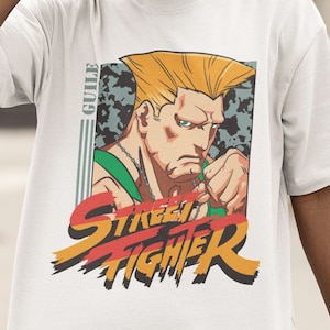 Puede incluir: Camiseta blanca con un estampado gráfico de un personaje de dibujos animados con cabello rubio y una camisa verde. El personaje está levantando un puño. El texto "Street Fighter" está impreso en letras rojas, amarillas y negras debajo del personaje. El texto "Guile" está impreso en letras negras a la izquierda del personaje.