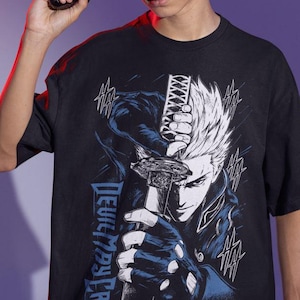 Può includere: Una t-shirt nera a maniche corte con una stampa grafica di un personaggio dai capelli bianchi a punta che impugna una katana. Il design include accenti blu e bianchi, il testo "DEVIL MAY CRY" e elementi testuali "HA" sparsi.
