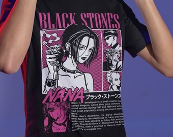 Puede incluir: Camiseta negra con un gráfico de manga de "Nana", incluyendo el texto "BLACK STONES" y caracteres japoneses. El diseño presenta a una mujer con un cigarrillo y otros personajes. La persona lleva unos auriculares.