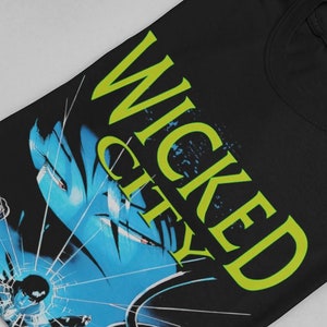 以下が含まれることがあります： 青い顔に割れたガラスの効果が施されたグラフィックプリントが施された黒いTシャツ。シャツには「WICKED CITY」というテキストが黄色で印刷されています。
