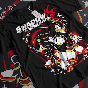Könnte beinhalten: Schwarzes T-Shirt mit einem Aufdruck von Shadow the Hedgehog aus der Sonic the Hedgehog Videospielserie. Der Aufdruck zeigt Shadow in einer dynamischen Pose mit einem rot-gelben Farbschema. Der Text "Shadow the Hedgehog" ist in Weiß über dem Aufdruck gedruckt.