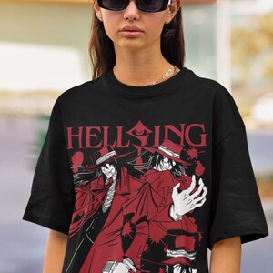 Może przedstawiać: Czarny t-shirt z czerwoną grafiką postaci z anime. Projekt zawiera postacie w czerwonych płaszczach i kapeluszach, z napisem "HELLSING" i "ALUCARD". Postacie trzymają broń.