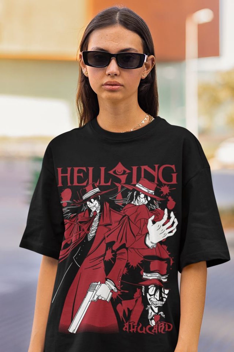 Może przedstawiać: Czarny t-shirt z czerwoną grafiką postaci z anime. Projekt zawiera postacie w czerwonych płaszczach i kapeluszach, z napisem "HELLSING" i "ALUCARD". Postacie trzymają broń.