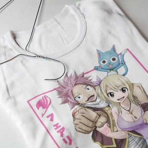 Peut inclure: T-shirt blanc avec un motif rose et blanc de trois personnages d'anime. Les personnages sont un garçon aux cheveux roses, une fille blonde et un chat bleu. Le texte "Fairy Tail" est écrit en japonais sur le t-shirt.