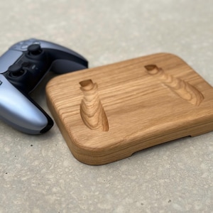 Op de afbeelding: Een houten standaard met twee gegraveerde sleuven om videogamecontrollers vast te houden. De standaard is gemaakt van lichtbruin hout en heeft een natuurlijke afwerking. Een grijze videogamecontroller ligt naast de standaard.
