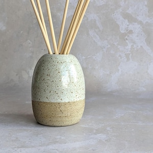 May include: A two-tone ceramic essential oil diffuser with light brown reeds. The vase has a speckled green upper section and a light brown base. The reeds are a light tan colour and are arranged in a fan shape from the top of the vase.