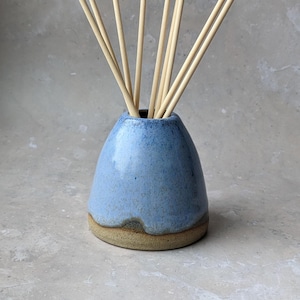 May include: A blue ceramic essential oil diffuser with a light brown base, filled with several light-coloured wooden reeds. The diffuser has a speckled finish and a rounded shape, ideal for home fragrance.