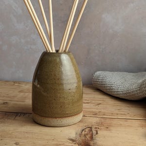 May include: A ceramic essential oil diffuser with light brown reeds. The vase is a muted olive green with a light tan base. The diffuser sits on a wooden surface, with a gray cloth in the background. Home decor.
