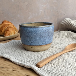 May include: A handcrafted ceramic bowl with a speckled blue exterior and a tan base. A small wooden spoon rests on a grey linen napkin. A croissant sits on a plate in the background.