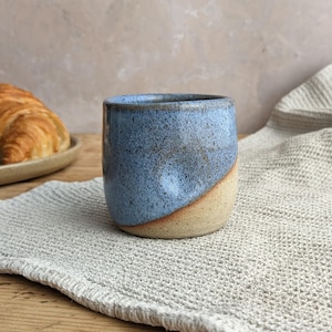 May include: A handmade ceramic mug with a blue and tan glaze. The mug has a rounded shape and a smooth finish. A croissant sits on a plate in the background. The mug is on a gray linen napkin on a wooden table.