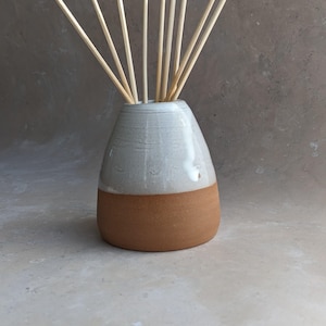 May include: A two-tone ceramic essential oil diffuser. The top half is light grey, the bottom is brown. Several wooden reeds are inserted into the opening, ready to diffuse fragrance. The vase has a smooth, glossy finish. The height is approximately 15 cm.