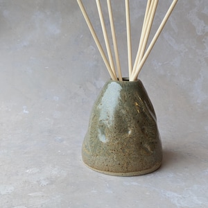 May include: A handcrafted, speckled ceramic essential oil diffuser in a muted green colour. The diffuser has a rounded shape and holds several light-coloured wooden reeds. The design is simple and natural, suitable for home fragrance.