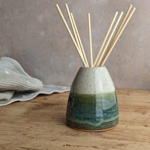 May include: A ceramic essential oil diffuser, featuring a speckled white top that fades into green and teal. The vase contains several light-coloured wooden reeds. The diffuser is placed on a wooden surface, creating a natural, spa-like atmosphere.