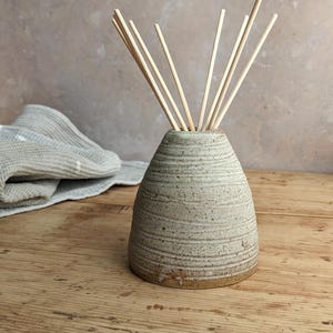 May include: A beige ceramic essential oil diffuser with a textured finish. The diffuser has a conical shape and contains wooden reeds. A folded grey and white towel rests on the wooden surface.