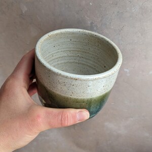 May include: Handmade ceramic mug with a speckled, off-white top, transitioning to a green and blue base. The mug has a smooth, rounded shape and a rustic, earthy aesthetic. Perfect for coffee, tea, or other beverages.