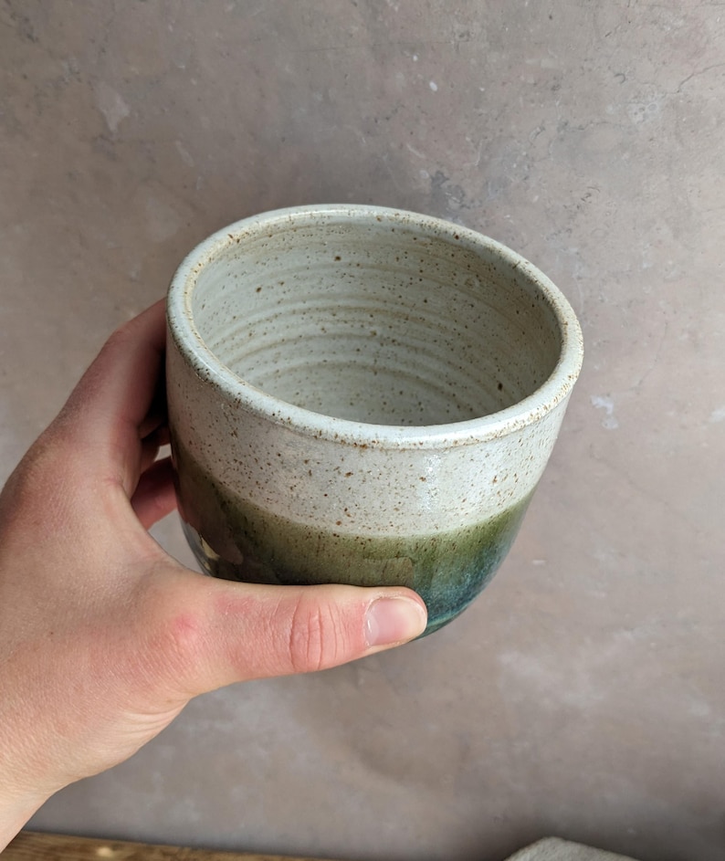 May include: Handmade ceramic mug with a speckled, off-white top, transitioning to a green and blue base. The mug has a smooth, rounded shape and a rustic, earthy aesthetic. Perfect for coffee, tea, or other beverages.
