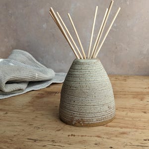 May include: A handmade, light-coloured ceramic essential oil diffuser with a conical shape and horizontal textured lines. The diffuser holds several wooden reeds, and sits on a wooden surface. A folded grey towel is in the background.