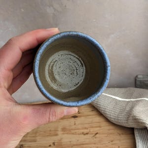 May include: A handmade ceramic cup with a blue rim and a speckled, earthy brown interior. The cup is held over a wooden surface, showcasing its rustic charm and unique design. The cup is a perfect size for a small beverage.
