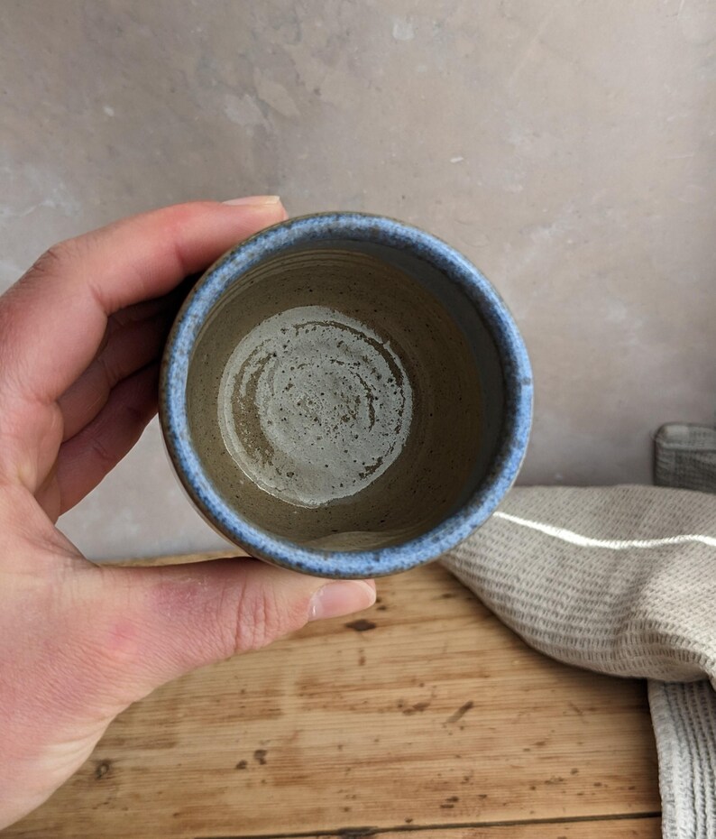 May include: A handmade ceramic cup with a blue rim and a speckled, earthy brown interior. The cup is held over a wooden surface, showcasing its rustic charm and unique design. The cup is a perfect size for a small beverage.