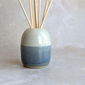 Könnte beinhalten: Ein zweifarbiges Keramik-Duftstäbchen-Diffusor mit hellblauer und weißer Glasur. Die Vase ist mit mehreren naturbelassenen Holzstäbchen gefüllt, die zur Duftverbreitung dienen. Ein dekoratives Wohnraumduft-Accessoire.