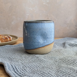 May include: A ceramic mug with a cylindrical shape and a rounded base. The upper portion features a speckled light blue glaze, while the lower portion remains an unglazed, textured beige clay. The rim is dark and smooth, contrasting with the body.