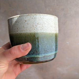 May include: A handmade ceramic cup with a speckled white top and a gradient of green and blue hues. The cup has a glossy finish and a rounded shape, perfect for holding beverages. The cup is held in a hand.