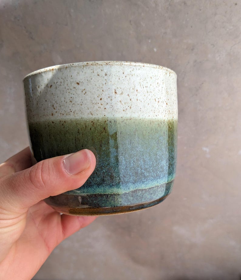 May include: A handmade ceramic cup with a speckled white top and a gradient of green and blue hues. The cup has a glossy finish and a rounded shape, perfect for holding beverages. The cup is held in a hand.